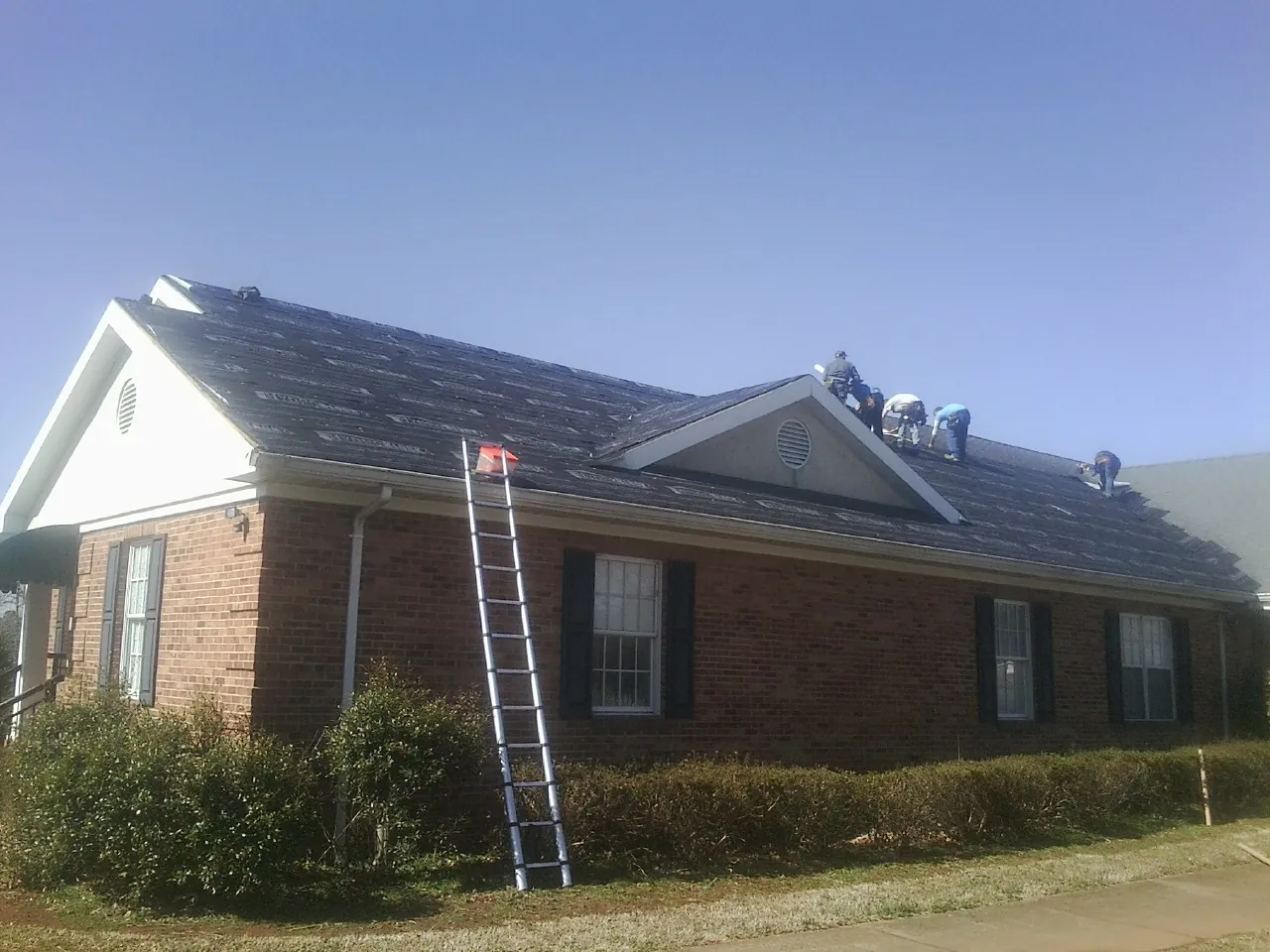 Crew tearing shingles off a brick ranch roof with a ladder set — Metal Roof Repair work by our crew in Midway Park