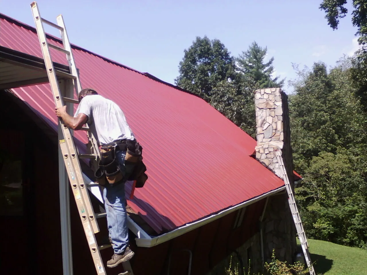 Roofer on a ladder beside a red metal roof and stone chimney — Soffit & Fascia Repair by Roofing Brothers in Midway Park